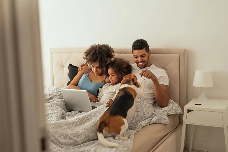 A family, mother, daughter, father, and family dog, a beagle, laying in bed watching a movie on a laptop.