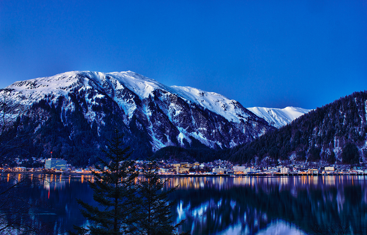 Winter view of Juneau, Alaska with snow covered mountains, city lights along the waterfront, and reflections on calm water at dusk.