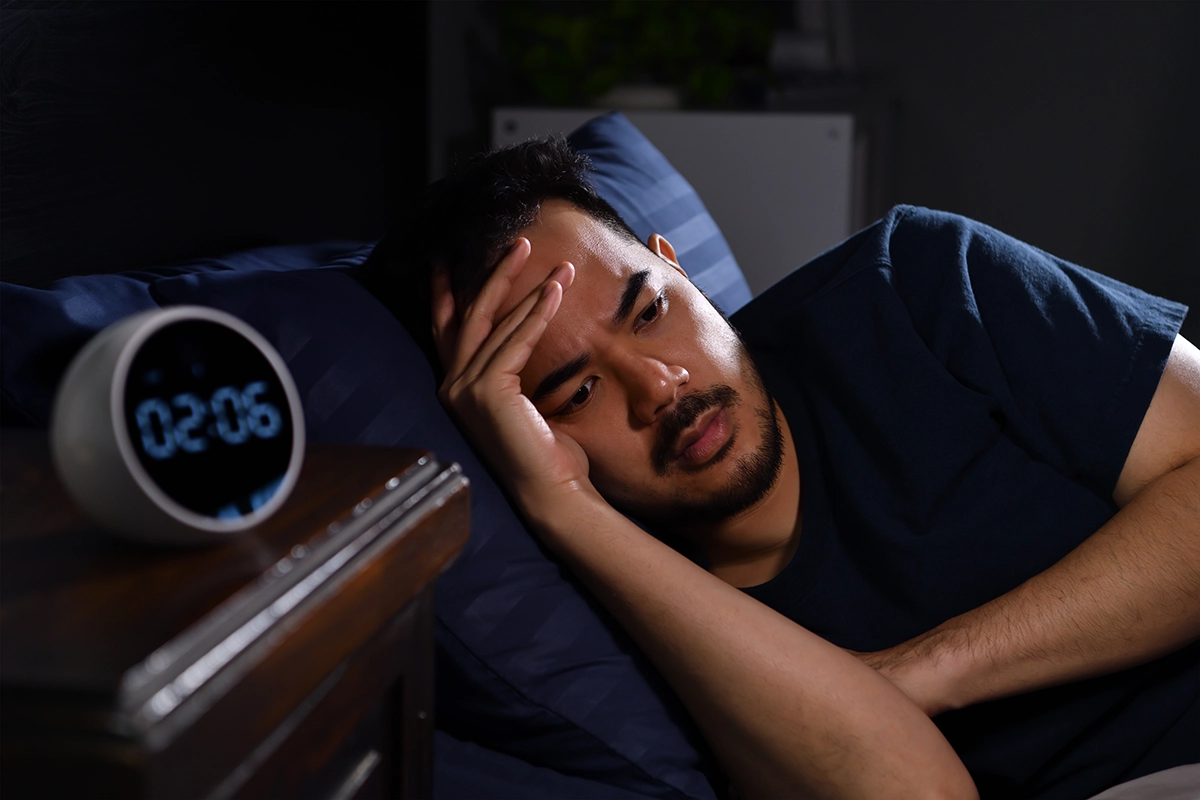 Man lying awake in bed at night, looking stressed while checking the clock at 2:06 AM.