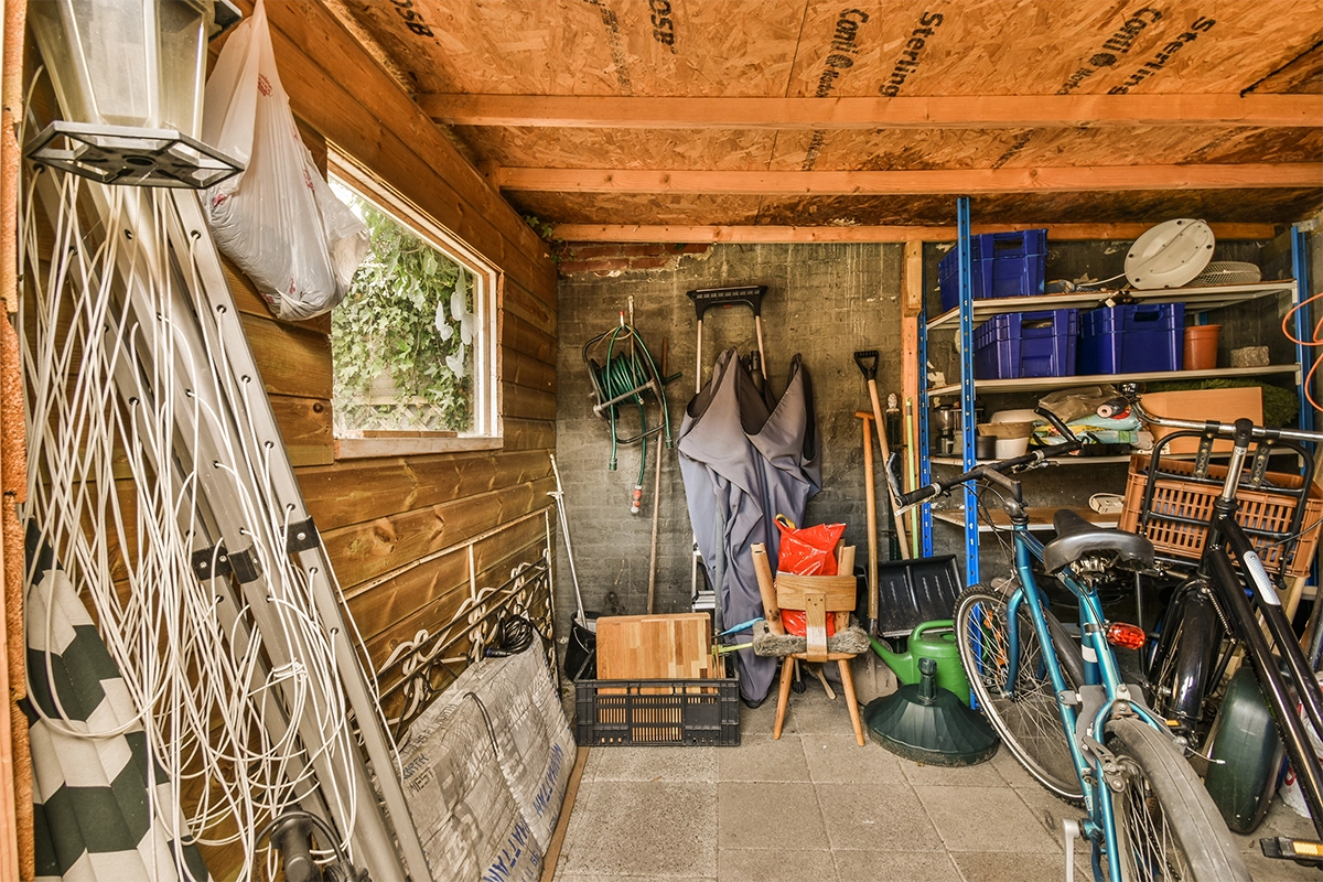 Cluttered storage shed filled with bicycles, tools, gardening supplies, and miscellaneous items.