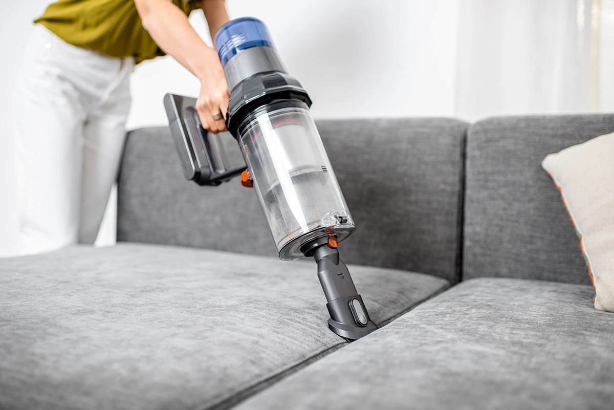 Person using a cordless vacuum cleaner to clean a gray couch, demonstrating home cleaning and pest prevention.