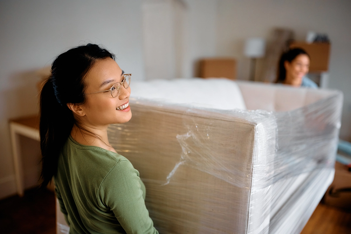 Two women smiling while carrying a wrapped sofa during a home move, representing safe furniture handling and clean moving practices.