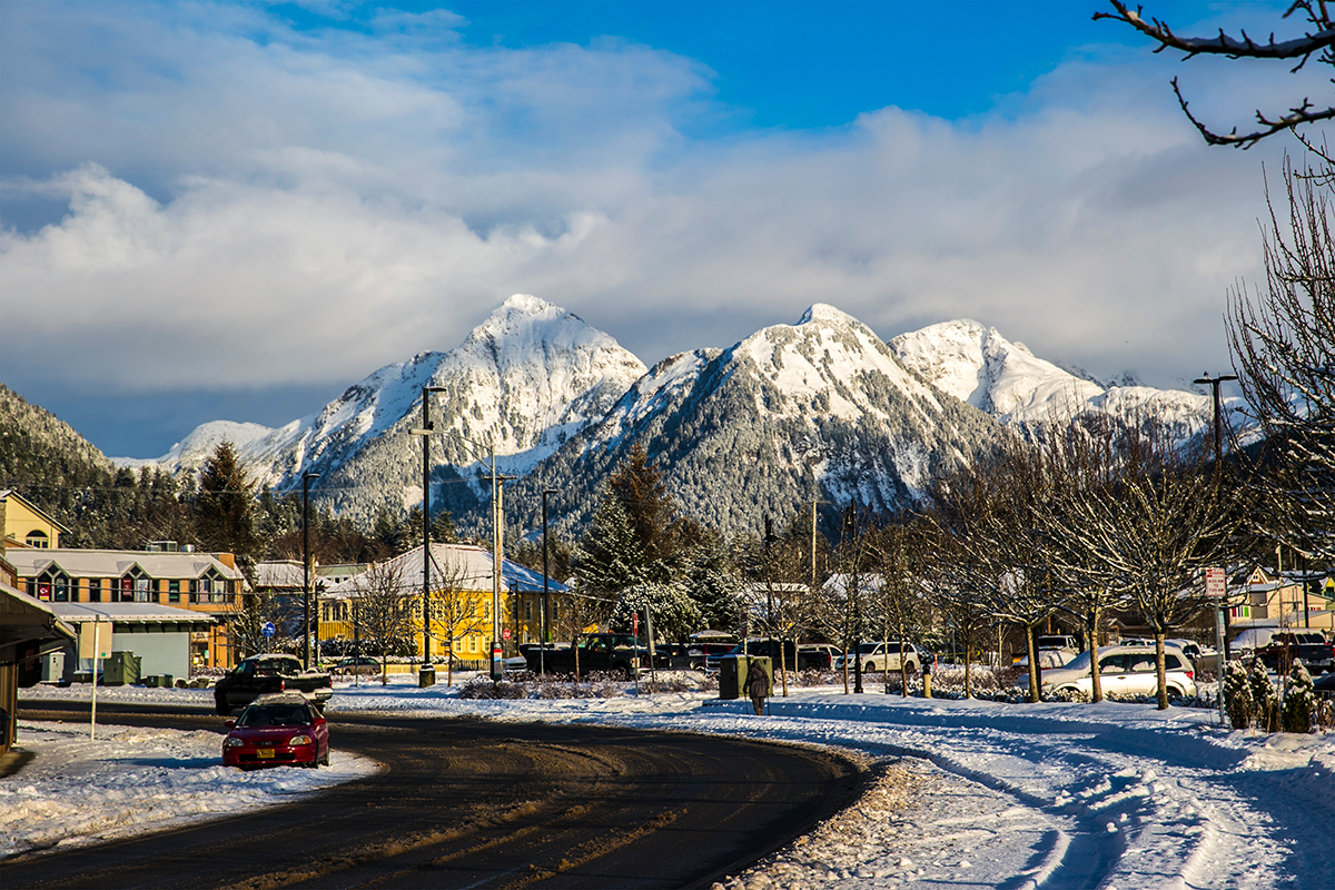 Winter street view in Sitka, Alaska with snow covered roads, parked cars, buildings, and snow capped mountains in the background.