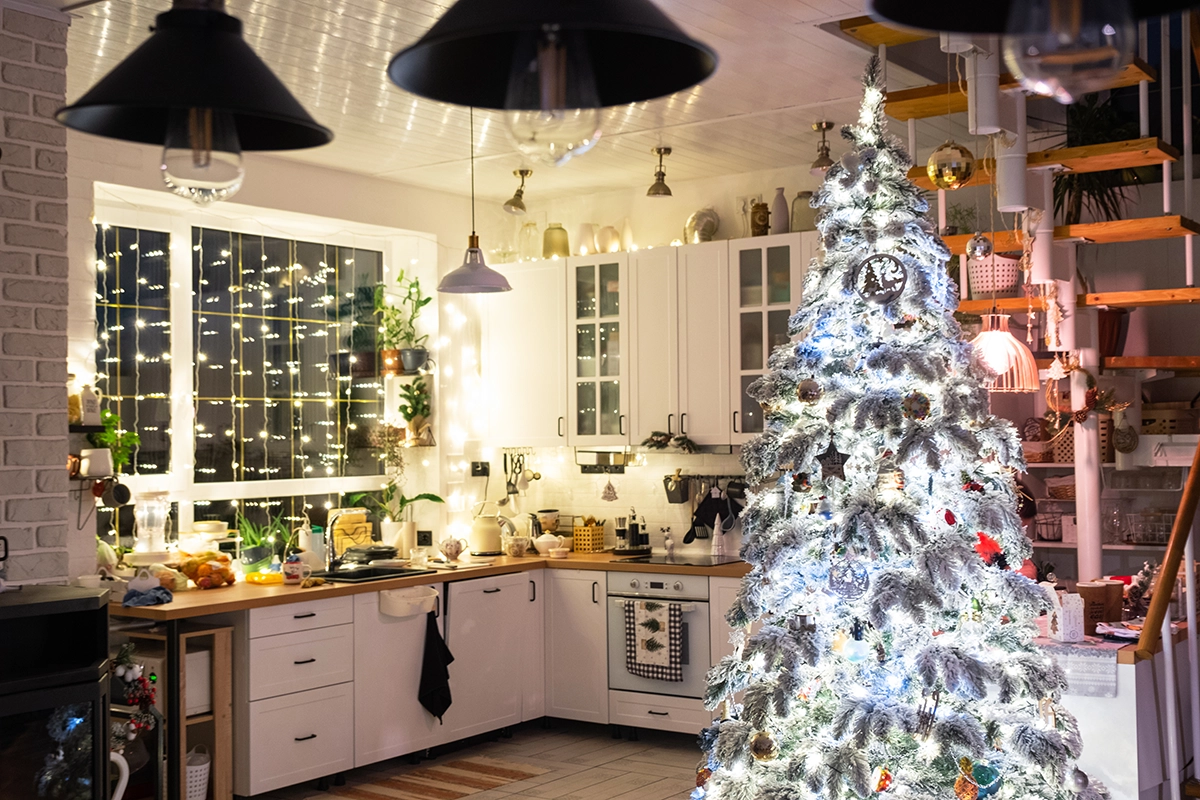 White kitchen decorated with Christmas lights and a bright, fully lit Christmas tree beside the counter.