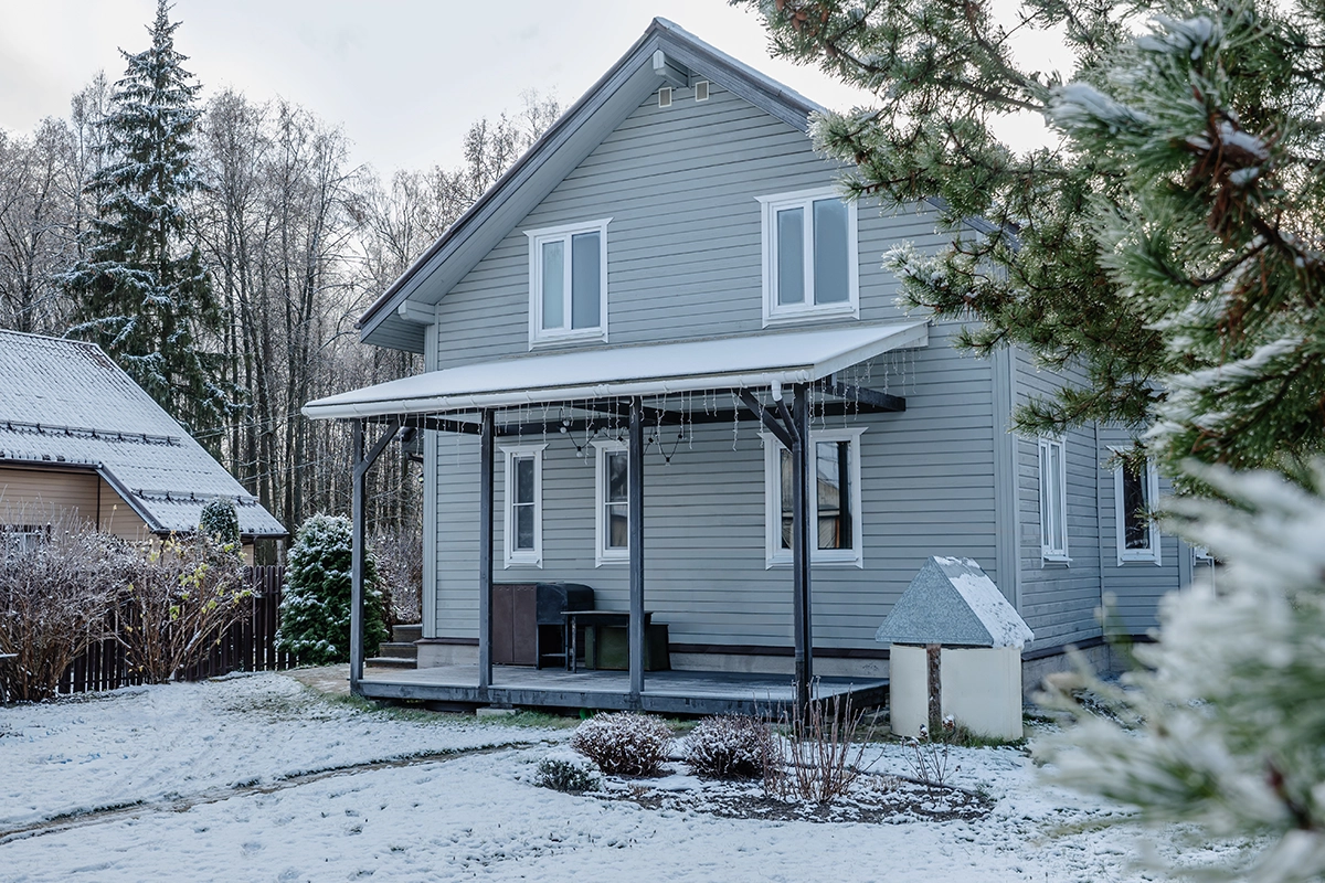 Gray two story house with a covered porch surrounded by snow covered ground, trees, and nearby buildings in winter.