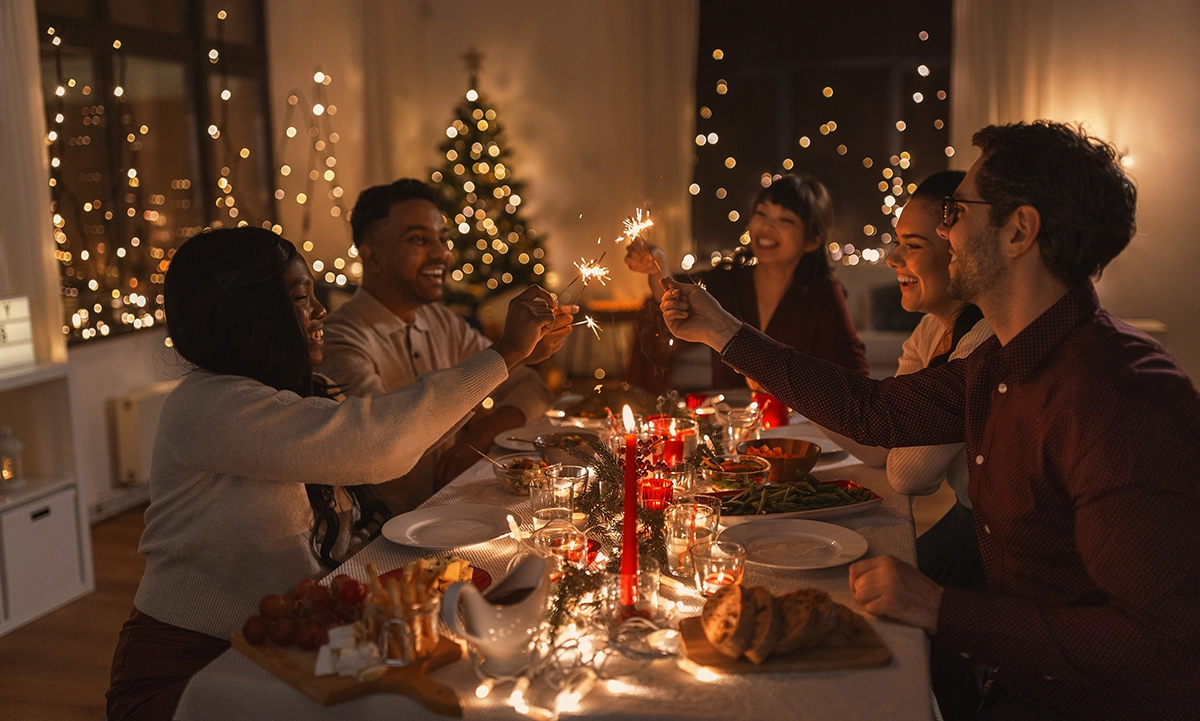 Group of friends celebrating at a festive dinner table while holding sparklers with holiday lights in the background.