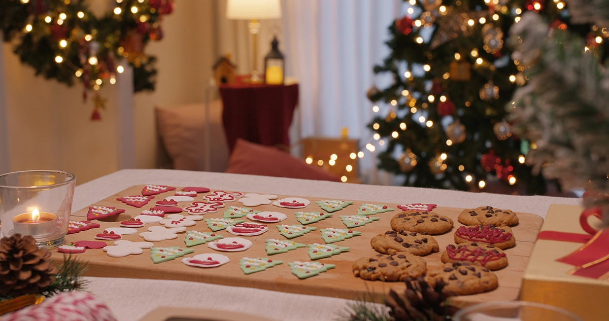Decorated Christmas cookies arranged on a wooden board with holiday lights, a wreath, and a Christmas tree glowing in the background.