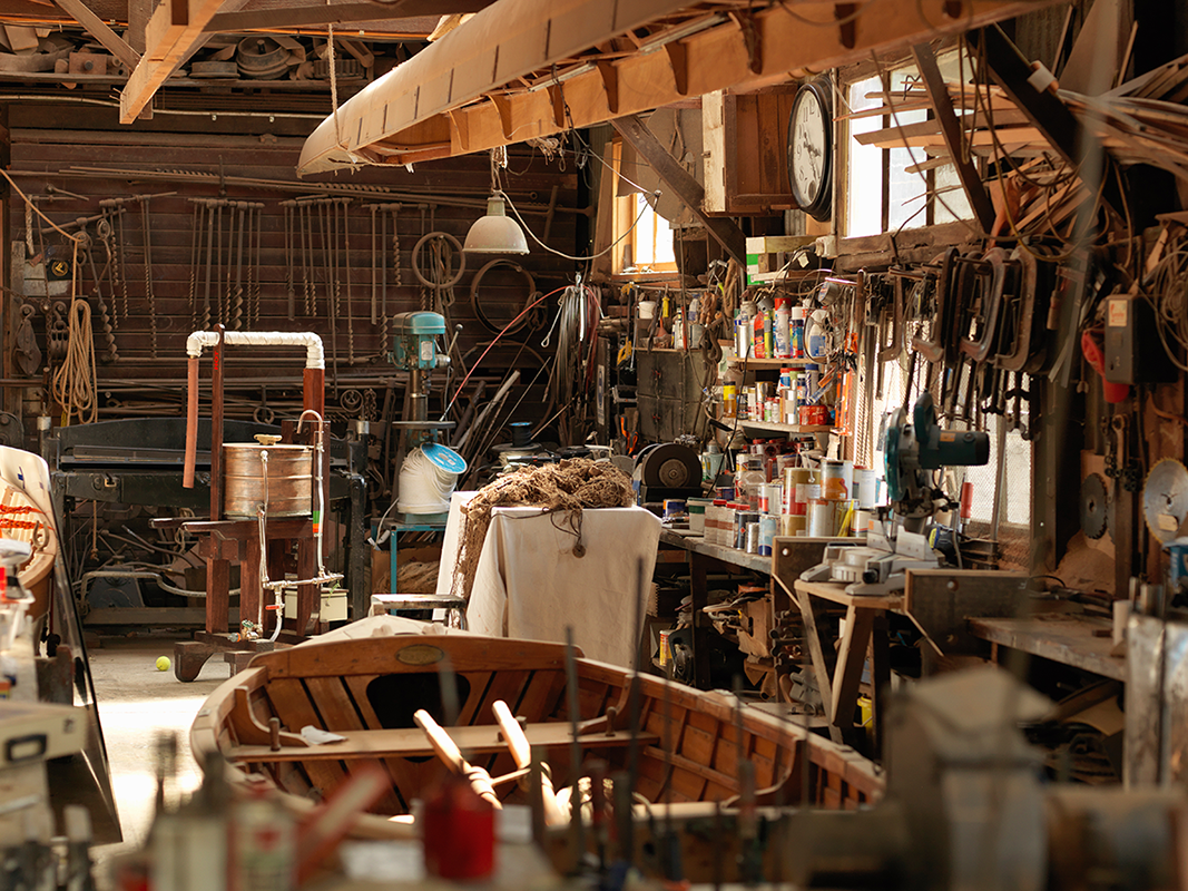 Cluttered workshop and garage interior filled with tools, supplies, shelving, and unfinished woodworking projects.