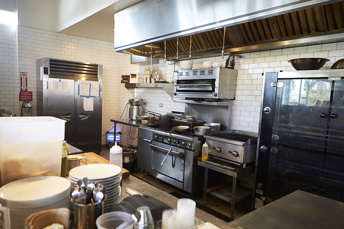 Commercial restaurant kitchen with stainless steel cooking equipment, prep surfaces, and ventilation hood.