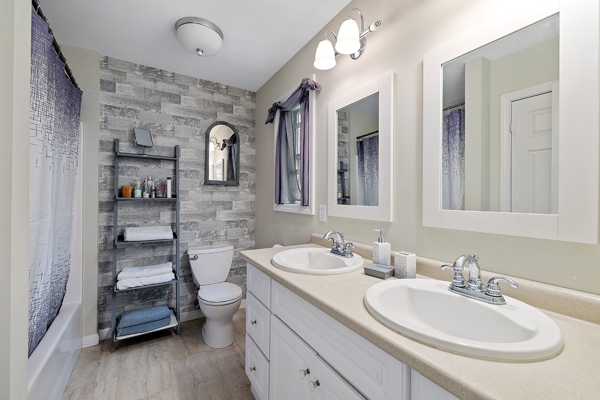 Bright bathroom with double sinks, large mirrors, a bathtub, shower curtain, toilet, and towel ladder against a gray accent wall.