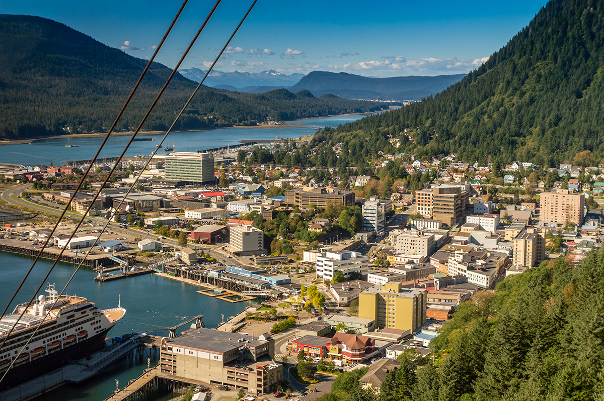 Aerial view of downtown Juneau, Alaska, with waterfront buildings, cruise ships, mountains, and forested hills.